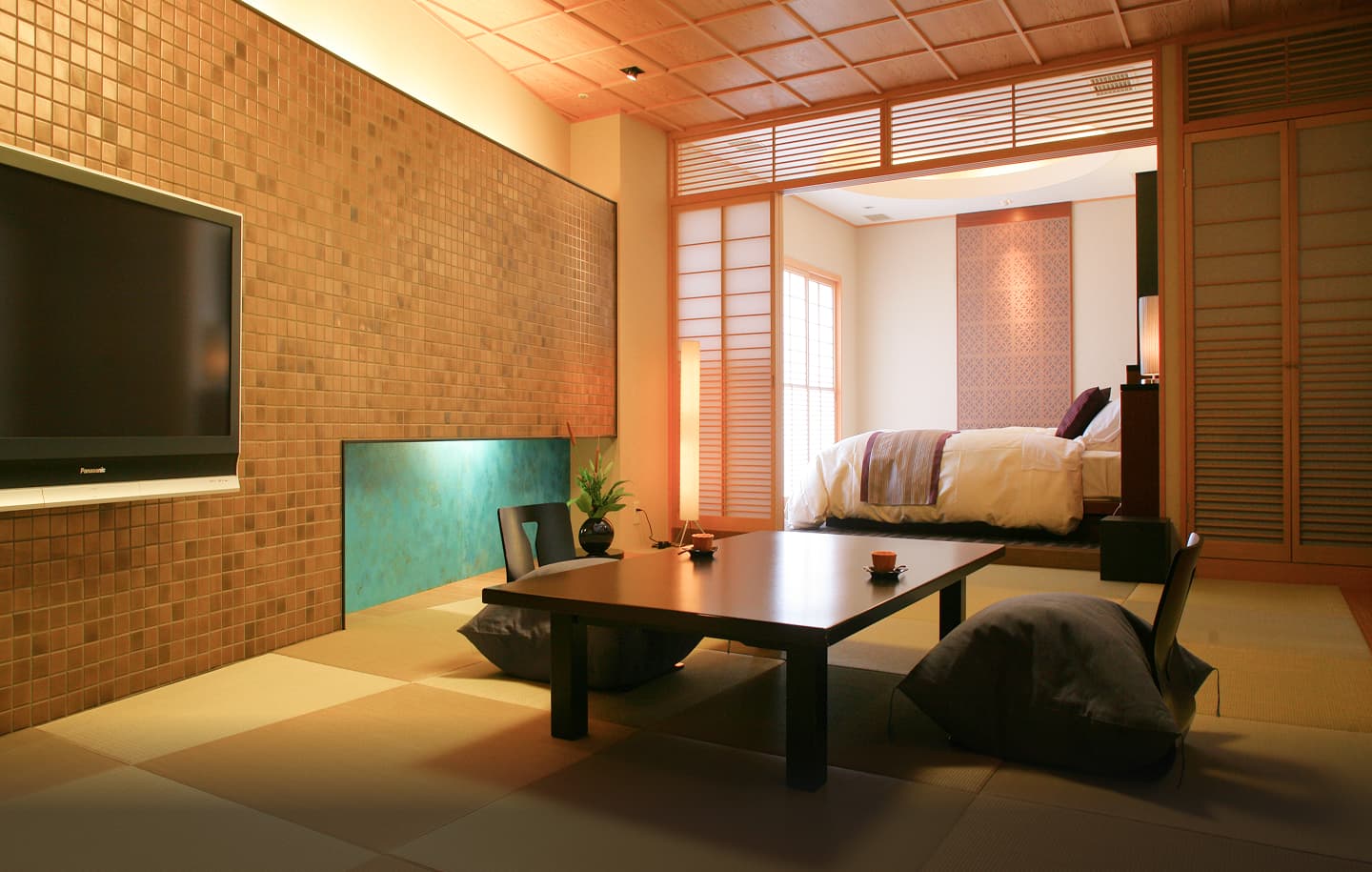 温かい照明に包まれた、広々としたバリアフリー対応の和洋室の全景。手前は畳敷きの空間で、中央にはカーブを描く木製の座卓が置かれ、その周りには座椅子やクッションが配置されている