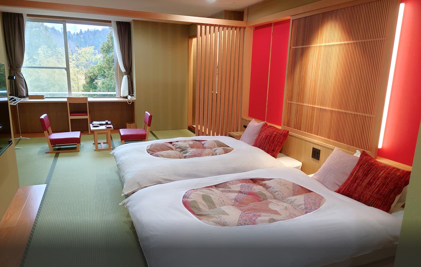 温かい間接照明に照らされた、広々とした和モダンな客室の全景。手前のリビングスペースは畳敷きで、中央には艶やかな黒い座卓が置かれ、その周りには黒い座布団と座椅子が配置されている。奥には木製の格子と障子戸で仕切られた寝室があり、2つのベッドが並び、清潔感のある白い布団が整えられている。