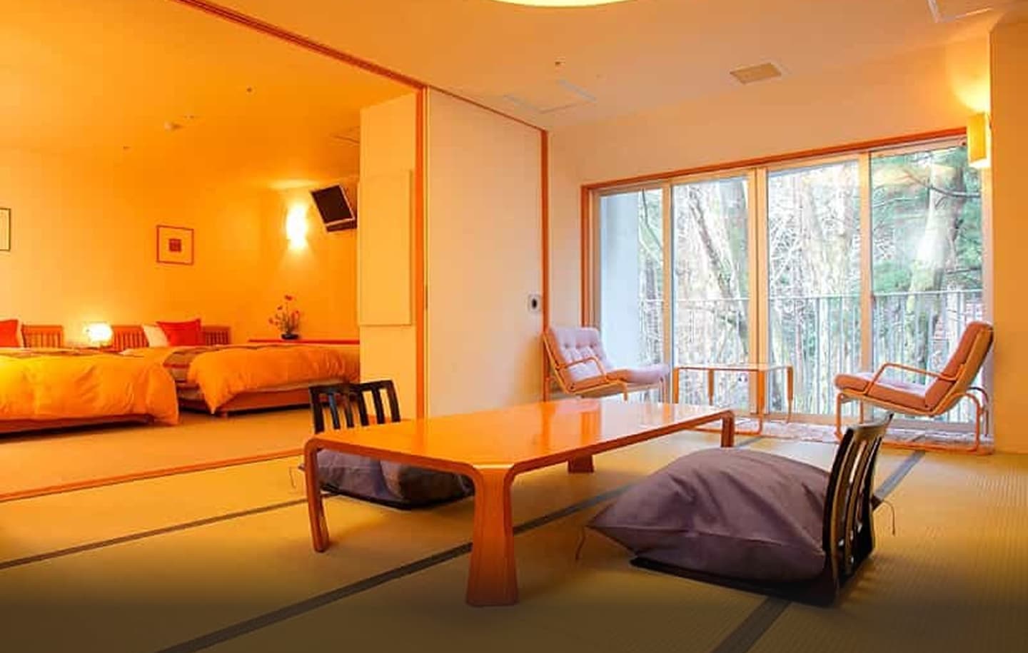 木目と畳の温もりが感じられる、広々とした和モダンな客室の全景。手前のリビングスペースは畳敷きで、中央には艶やかな黒い座卓が置かれ、その周りには座椅子が配置されている。奥には引き戸で仕切られた寝室があり、広々としたベッドには落ち着いた色合いの寝具が整えられている。