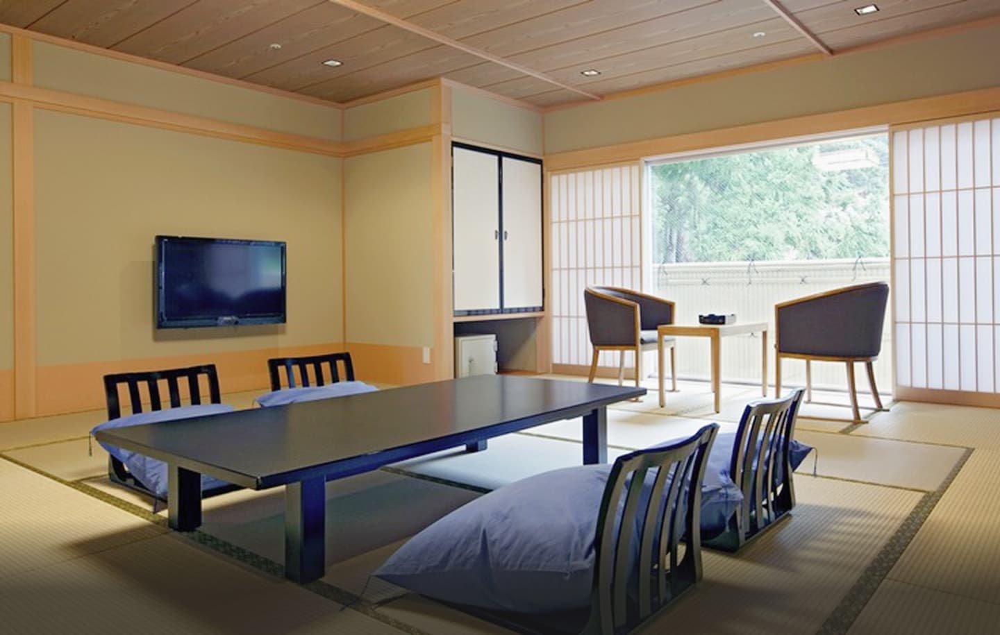 伝統的な和の趣と現代的な快適さが融合した、広々とした旅館の和室の全景。手前には畳が敷かれ、中央には黒い大きな座卓が置かれ、その周りには座椅子と座布団が整然と並べられている。