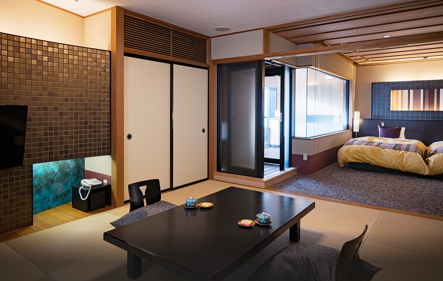 落ち着いた色調で統一された、広々とした旅館の和洋室の全景。手前の和室部分には、墨色の座卓が置かれ、その周りには座布団と座椅子が配置されている。奥には、引き戸で仕切られた寝室があり、2つのベッドには暖色系の布団がかけられ、壁には抽象画のような飾りが掛けられている。